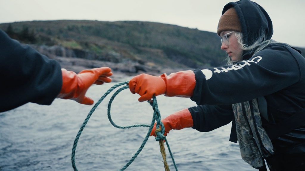A poignant moment of teamwork and tradition as Alicia Warford gracefully hands the buoy's rope to her father aboard their fishing boat, captured in mesmerizing slow motion. Against the backdrop of Newfoundland's rugged coastline, Alicia and her father exemplify the synergy and camaraderie essential to the fishing life.