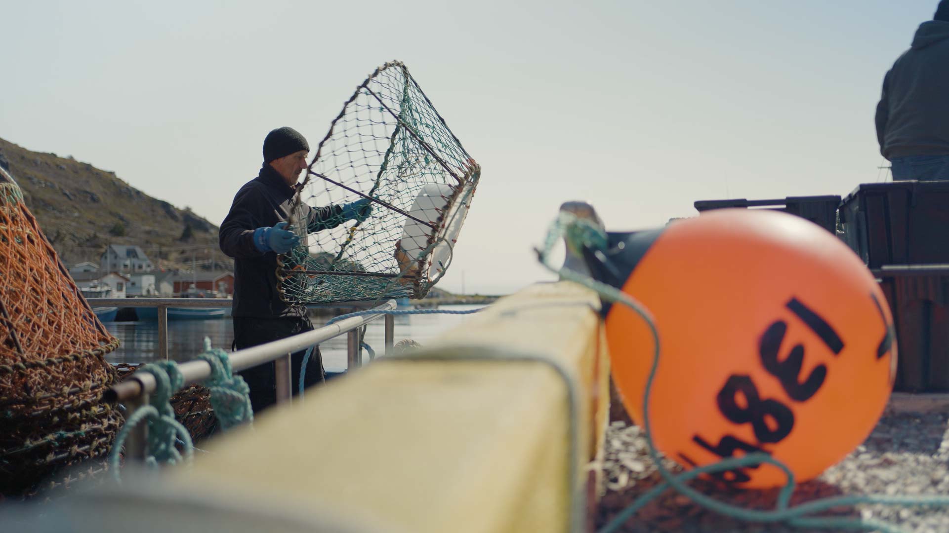 Des membres de l'équipage chargent des casiers à crabe à la main sur le bateau de pêche dans le port, avec une bouée visible au premier plan.